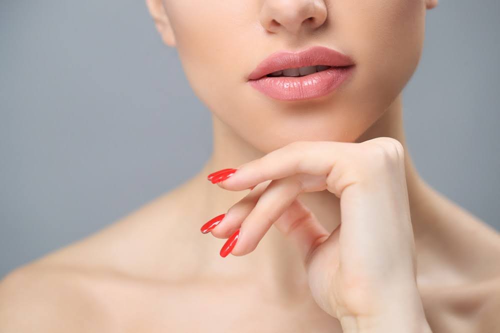 Close-up of a woman's lips and hand, with natural pink lipstick and red manicured nails, touching her chin