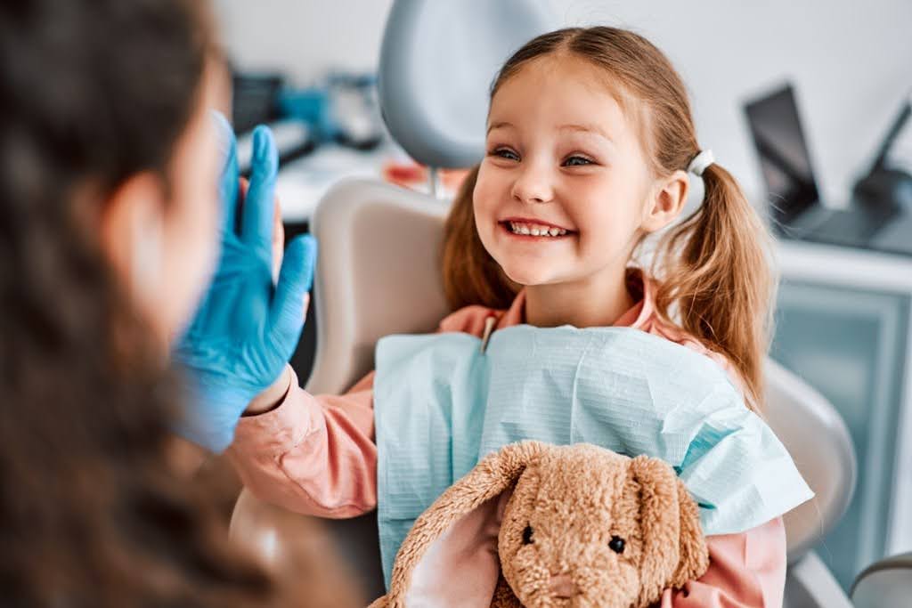  A smiling young girl in a dental chair high-fives her dentist while holding a plush bunny.