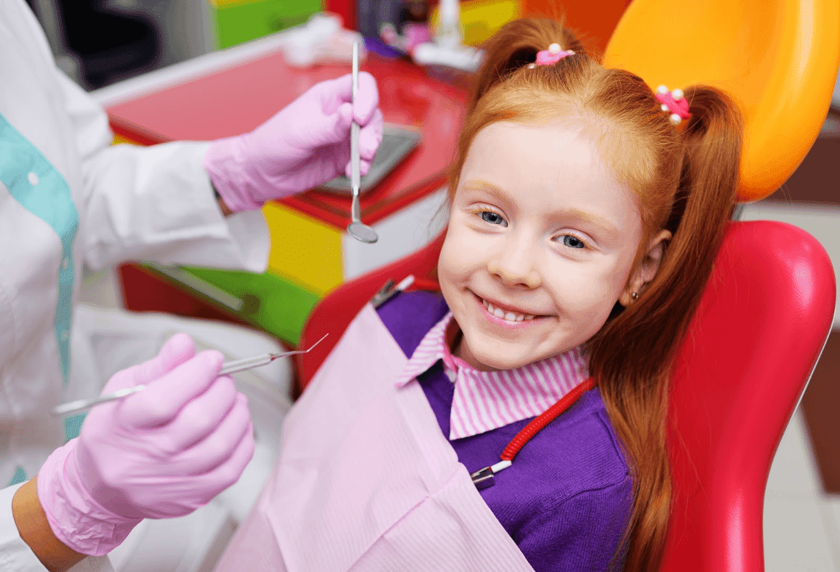   A smiling young girl with red hair sits in a dental chair as a dentist in pink gloves prepares for her checkup.