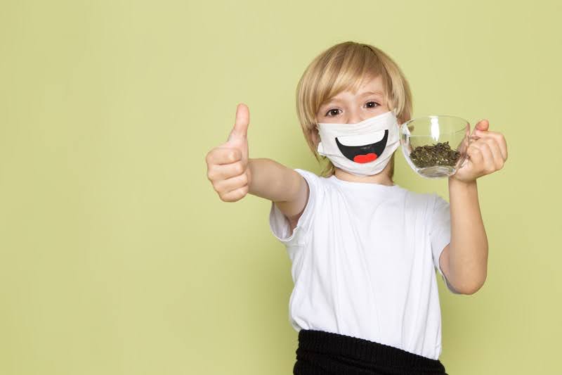  A kid with a smiling dust mask giving thumbs while holding a cup, waiting for his metal crown placement